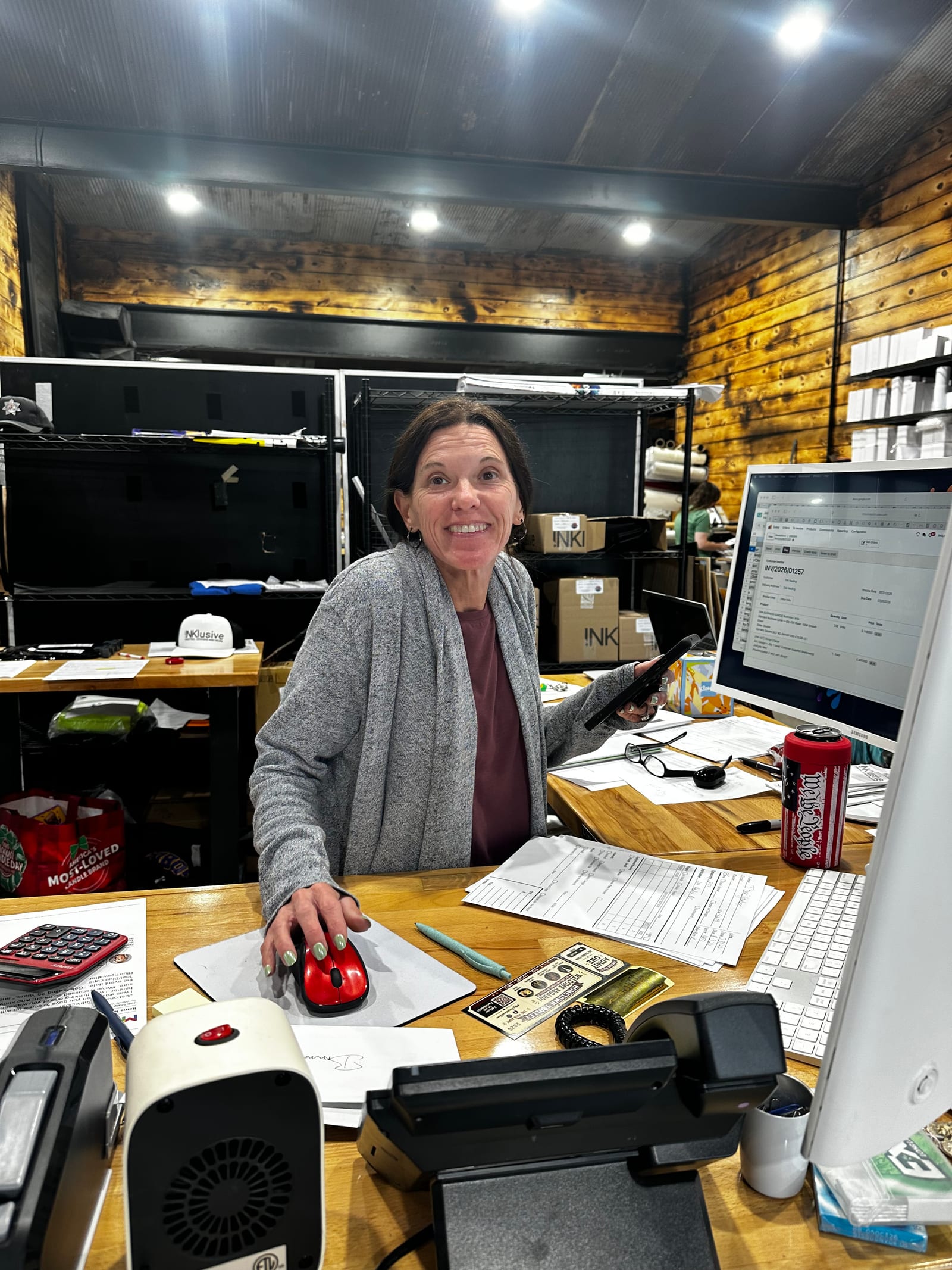 A staff member at INKlusive's front counter, smiling toward the camera while working at the order desk. An order form is open on the monitor beside her, with printed paperwork, a card reader and shop supplies across the wooden counter.