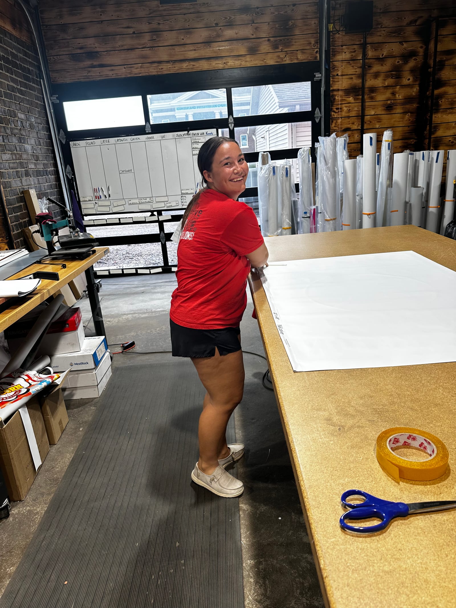 A staff member in a red INKlusive shirt at the wide-format work table, looking back from a large white printed sheet laid out for trimming. Rolls of wide-format material are racked behind her, beside the production schedule board and the open bay door.