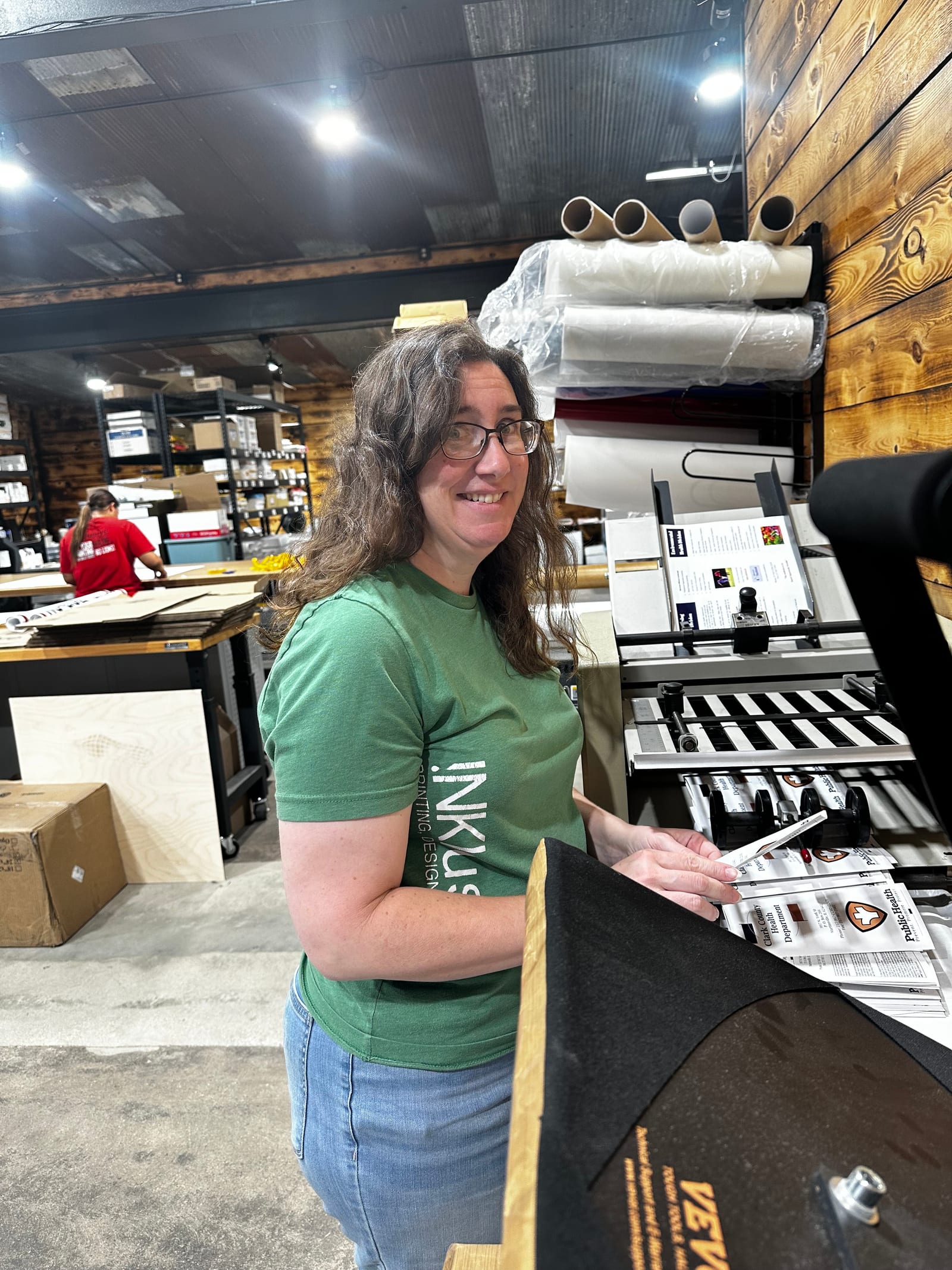 A staff member in a green INKlusive shirt working printed sheets through the finishing equipment, with rolls of laminate stored overhead and a colleague at a bench further down the production floor.