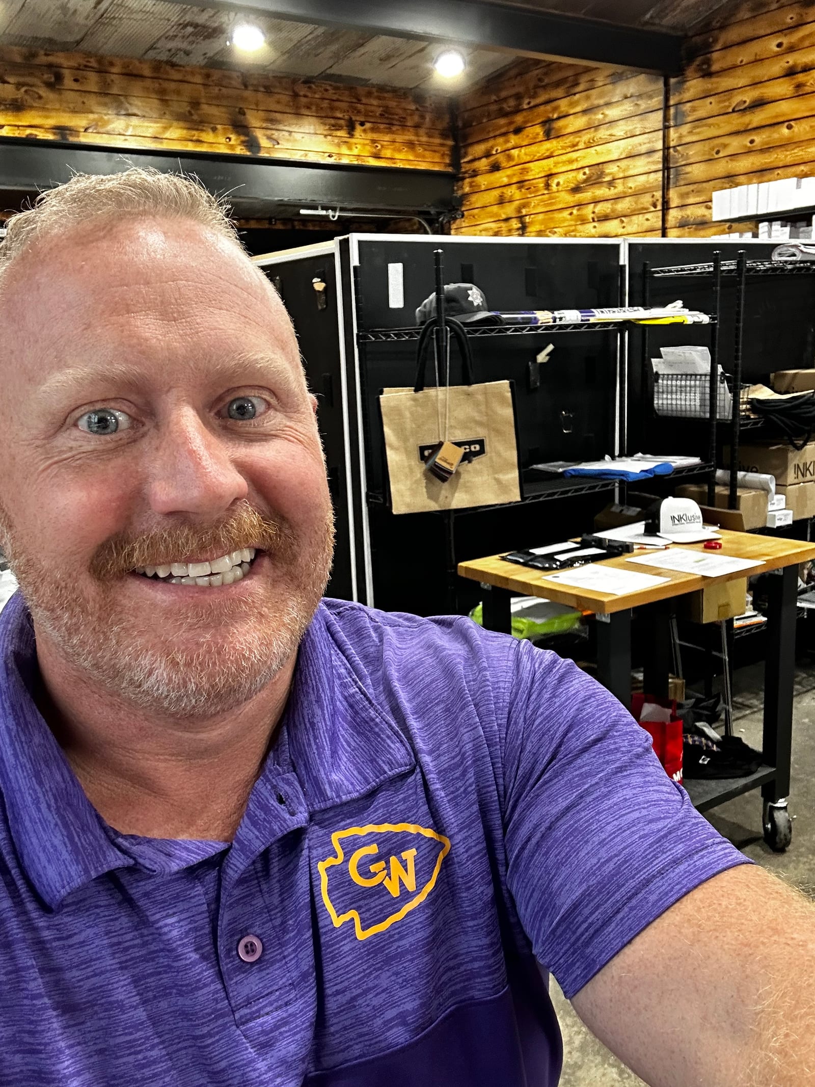 A close candid on the INKlusive production floor. On the shelving behind are branded tote bags, a cap, and a work table of printed materials.