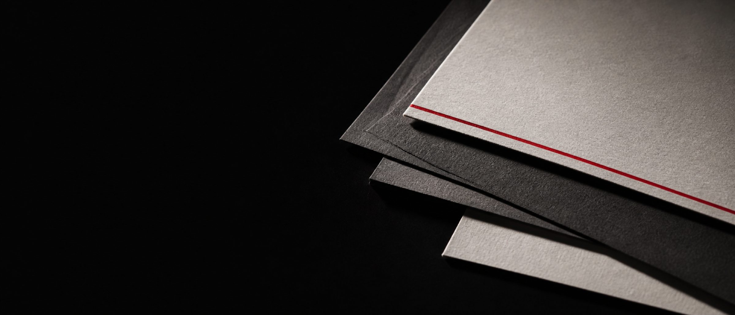 Two dark grey sheets of uncoated stock overlapping on a black surface, lit hard from one side, with a thin red printed line along the edge of the top sheet.