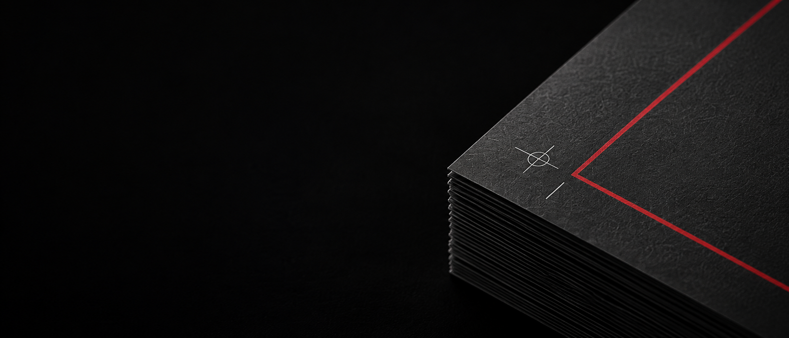 A stack of black printed cards seen edge-on against a black background, the top card carrying a thin red rule and a registration target, with open space to the left.
