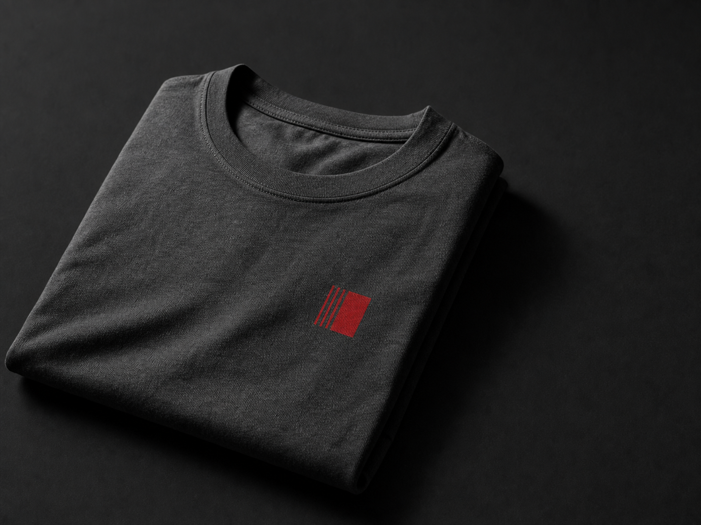 A folded charcoal cotton t-shirt on a black surface, lit so the jersey weave is visible, with a small red printed mark on the chest.