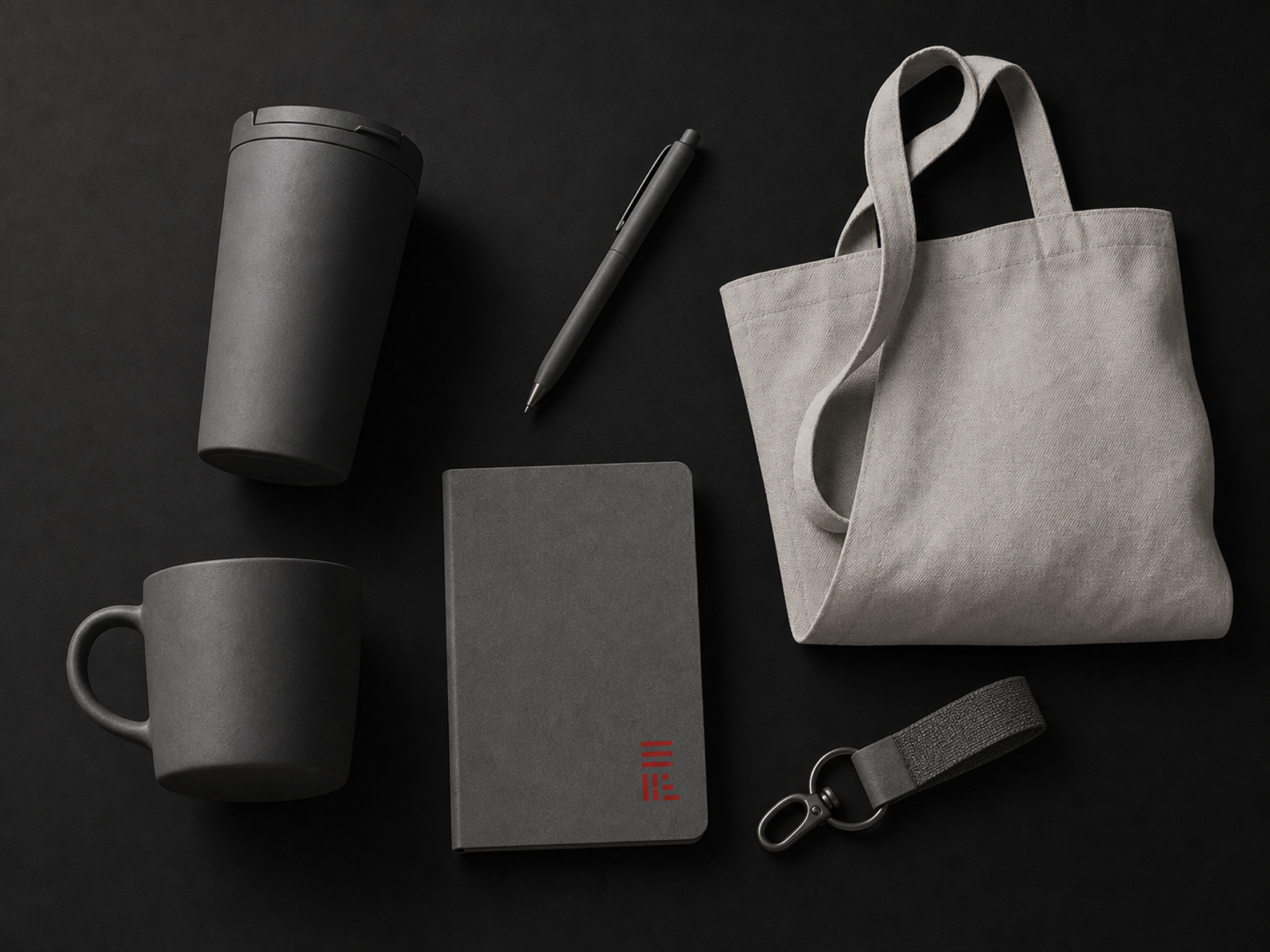 Six branded items on a black surface: an insulated tumbler, a pen, a canvas tote, a ceramic mug, a notebook carrying a small red mark, and a keyring.